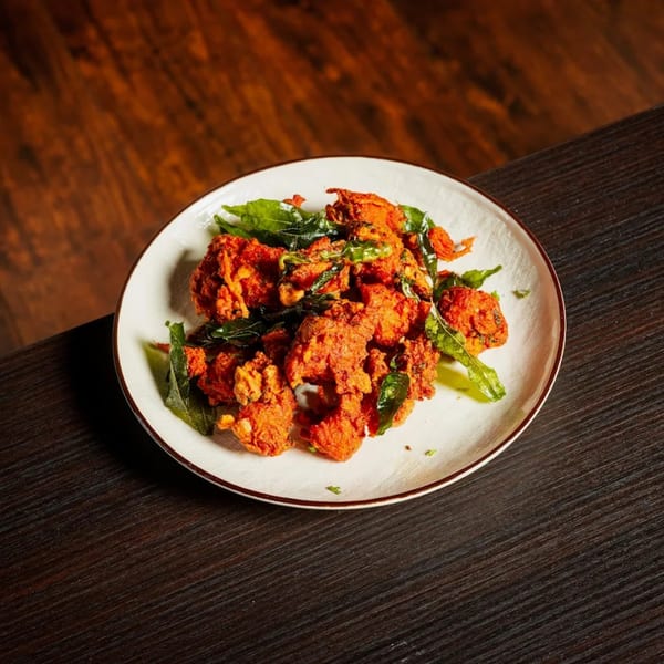A white plate filled with vibrant orange fried chicken pakora and green curry leaves on a dark wooden table.