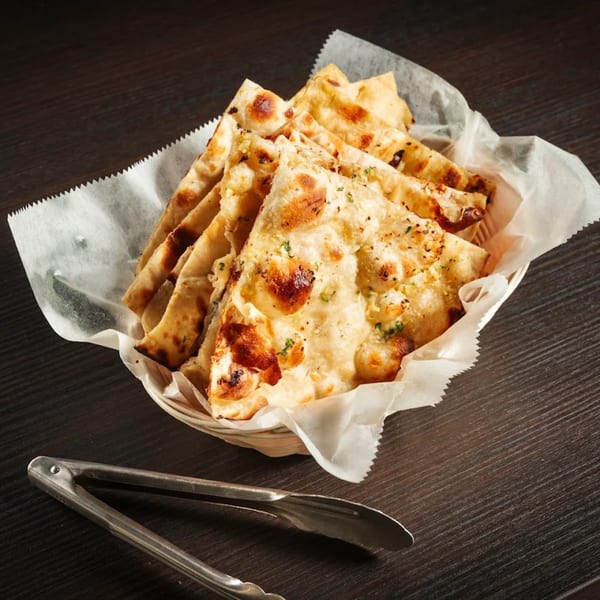 Basket of golden-brown Indian naan bread, cut into triangles, with metal tongs on a dark wooden table.