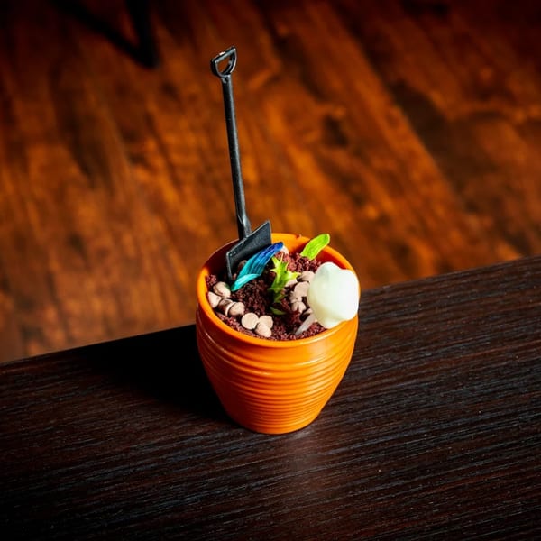 A dessert in an orange pot, resembling soil with chocolate chips, a white mushroom, and a small shovel.